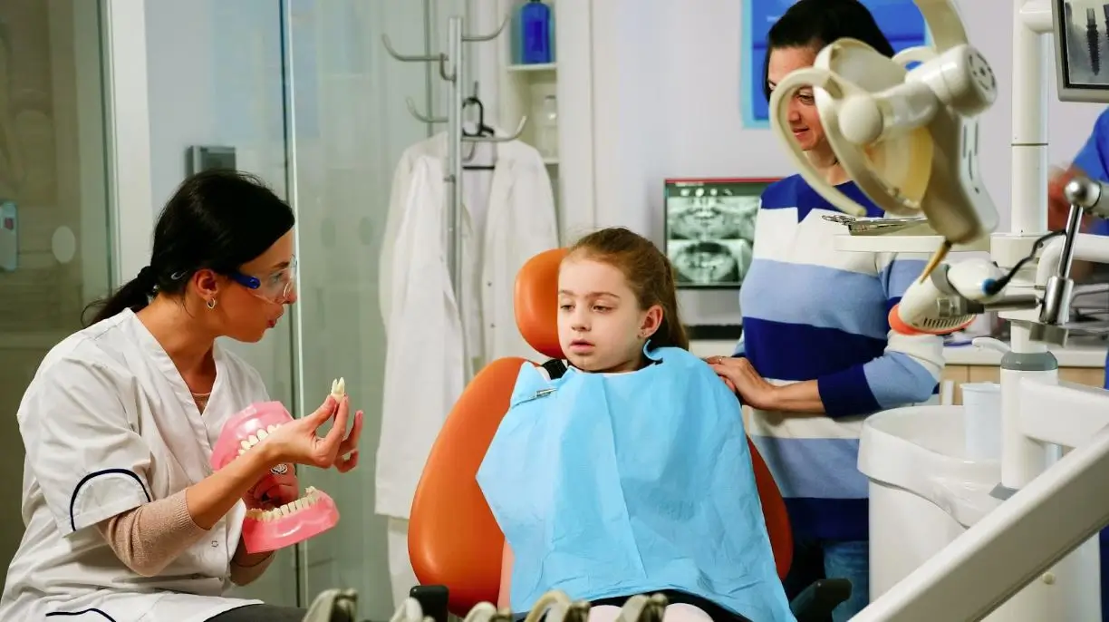 A dentist explains dental hygiene techniques to a young patient using a dental model while the child's parent stands nearby.