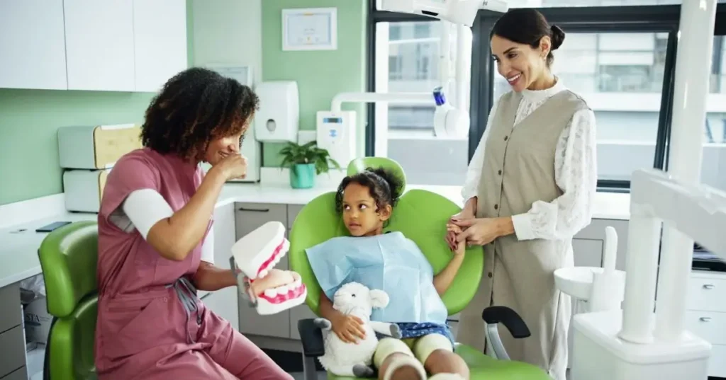 A dentist uses a dental model to demonstrate oral hygiene techniques to a young patient holding a stuffed toy while the child's parent stands nearby.