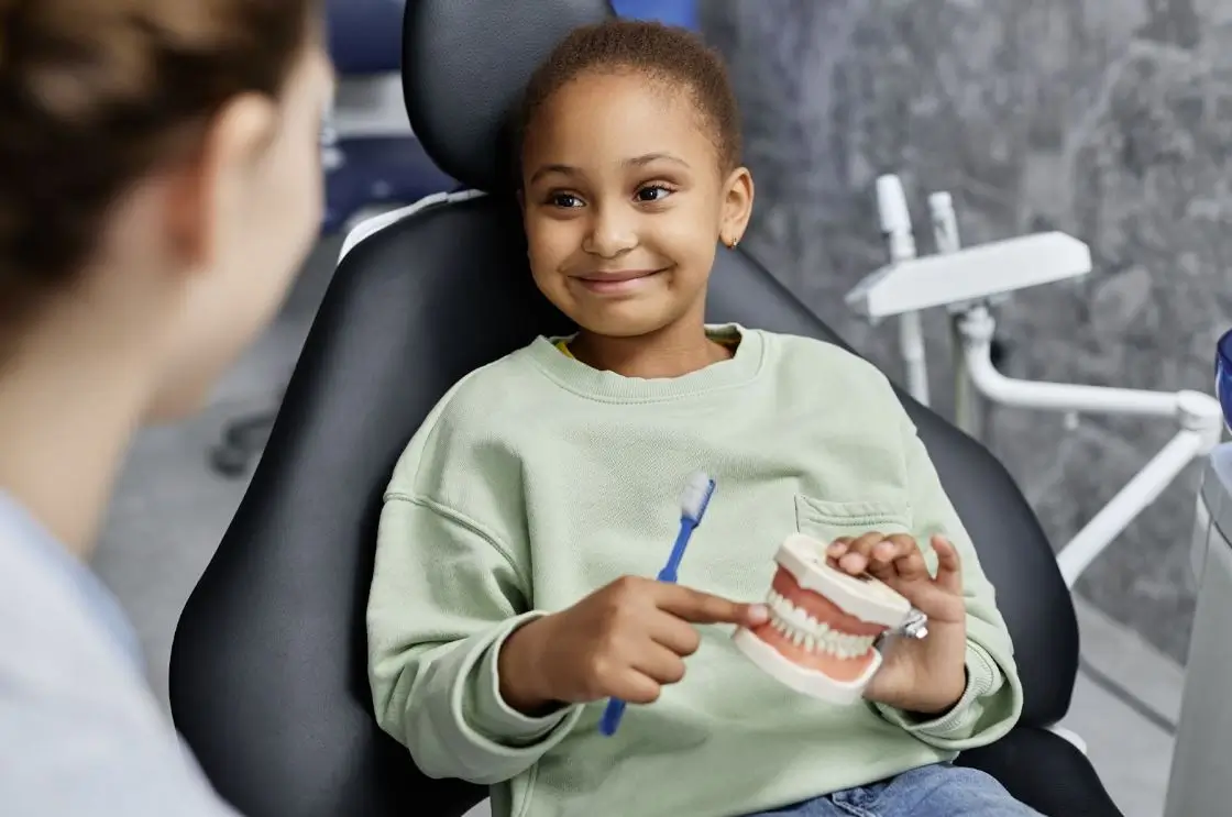 A young girl in a dental exam chair is holding a model of teeth and a toothbrush.