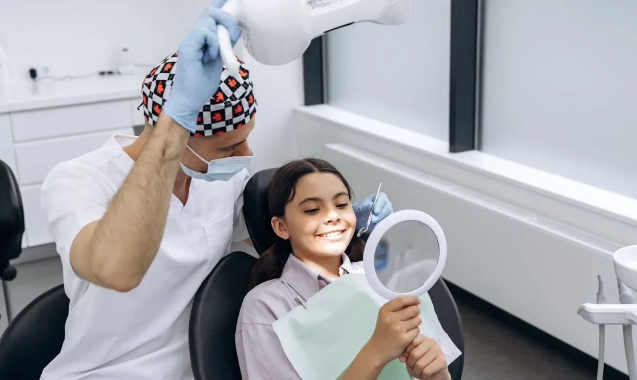 A young girl in an exam chair looking at her smile in a mirror next to a male dentist.