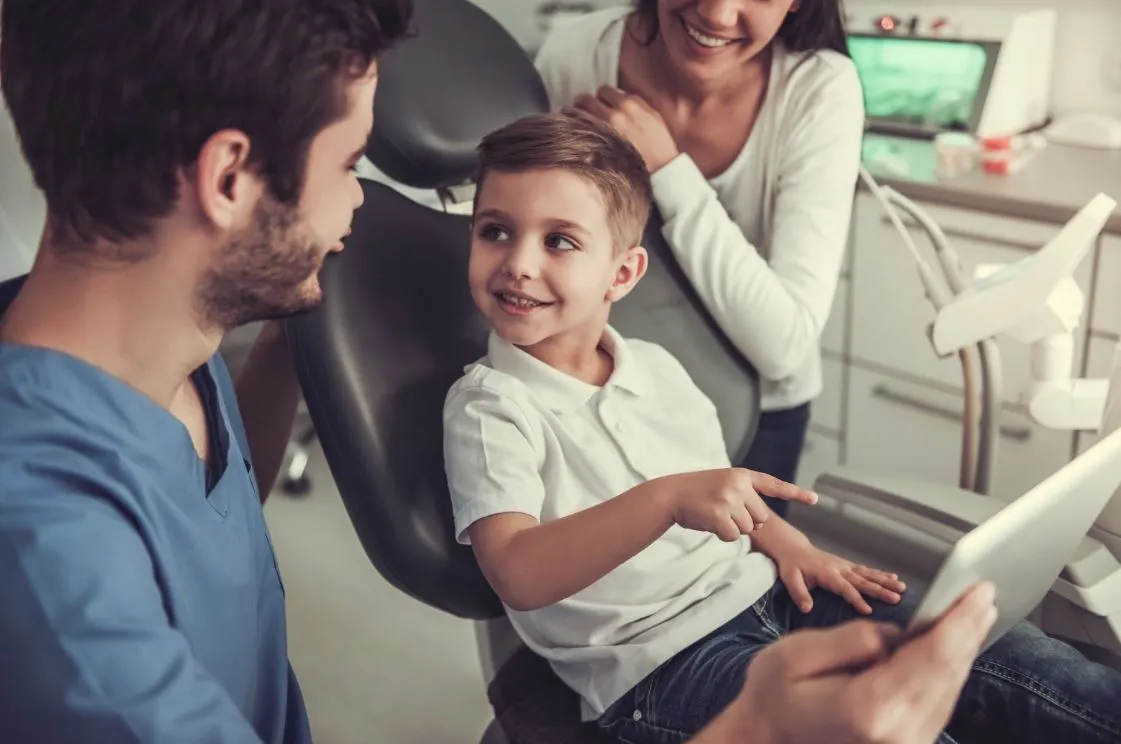 Woman standing beside a young boy in an exam chair beside a male dentist.