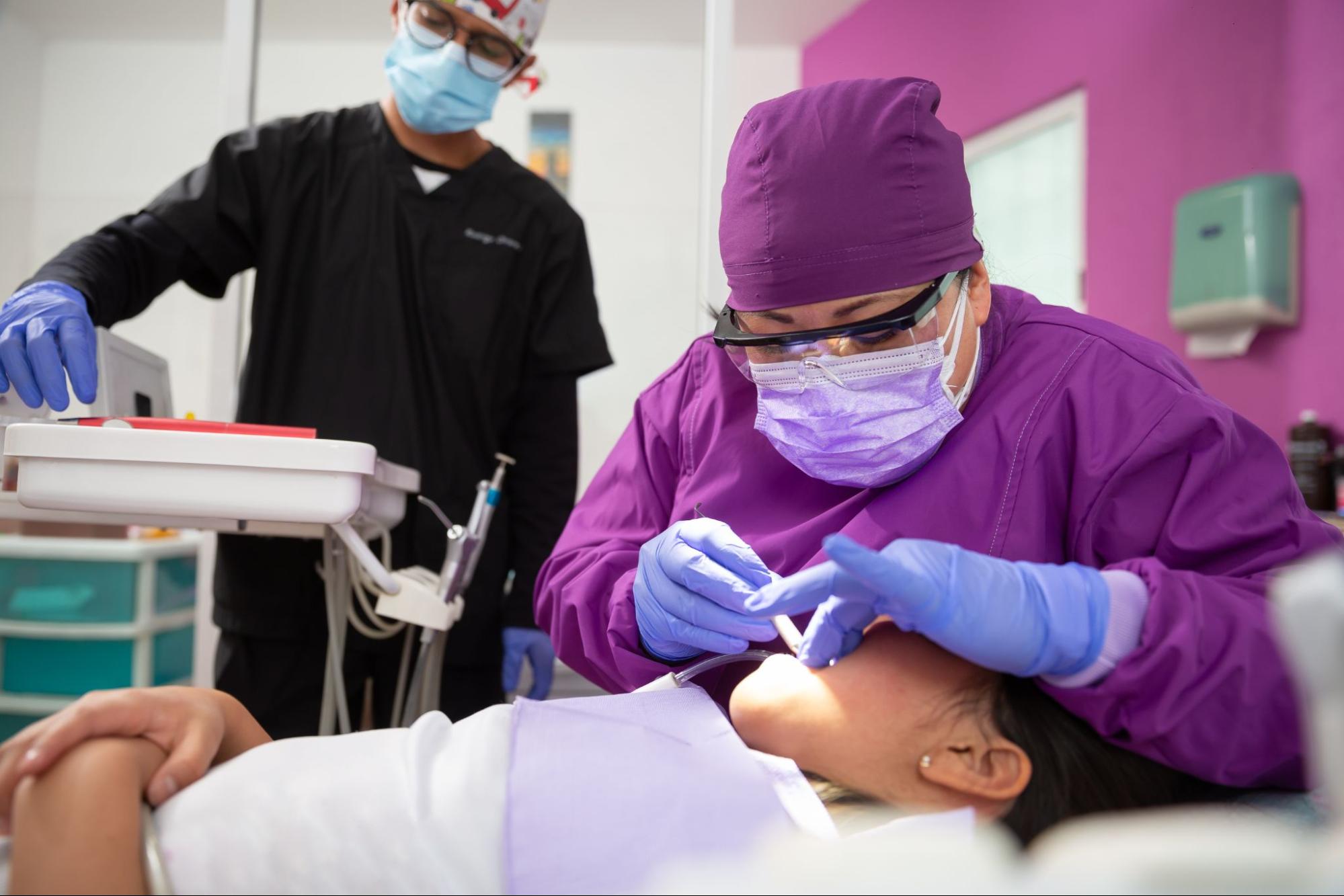 A lady dentist wearing a magenta medical uniform performs teeth cleaning to a patient while her assistant in the background organizes the dental tools. 